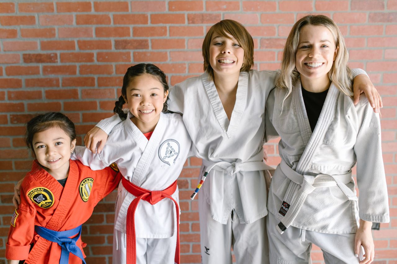 Happy children in taekwondo uniforms and belts pose against a brick wall, showcasing diversity and teamwork in martial arts.